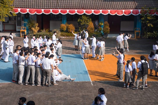 A large group of students in school uniforms, mostly in white shirts, are gathered outdoors in a school courtyard. Some are taking selfies while standing on colorfully painted sections of the ground. Others are mingling, and some are sitting on the ground. The building in the background has a tiled roof and is decorated with red and white bunting.