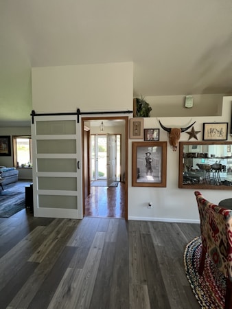 The image features an interior view of a home with modern decor. There is a sliding barn door to the left, leading into an entryway with natural light coming through the front door. The walls are adorned with artwork and decorations, including framed pictures and a mounted set of horns. A portion of the dining area is visible, featuring a patterned chair and reflective surfaces.
