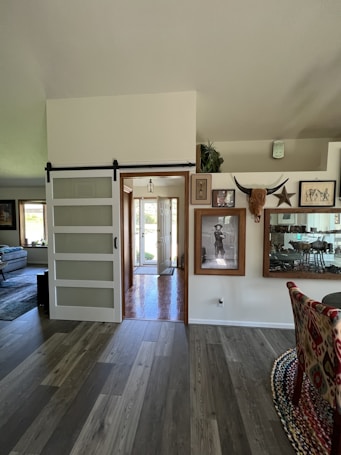 The image features an interior view of a home with modern decor. There is a sliding barn door to the left, leading into an entryway with natural light coming through the front door. The walls are adorned with artwork and decorations, including framed pictures and a mounted set of horns. A portion of the dining area is visible, featuring a patterned chair and reflective surfaces.