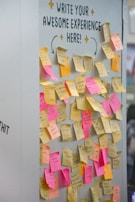 A wall is covered with numerous sticky notes in various colors such as pink, yellow, and orange. Above the notes, there is a message encouraging people to share their experiences. The sticky notes are filled with handwritten messages, some legible with phrases and doodles.