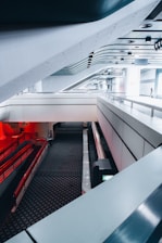 A modern architectural interior featuring a combination of sleek lines, metallic surfaces, and a multi-level design. The space is characterized by its futuristic aesthetic, with curved structures and a ceiling composed of grids and vents. Red lighting accents and patterned flooring create a dynamic visual contrast.