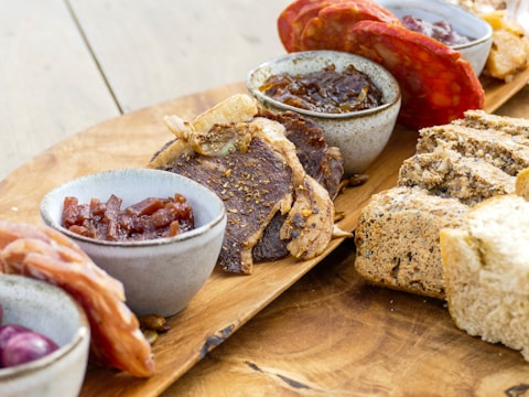 A wooden platter is arranged with a variety of cured meats, artisan breads, and small bowls containing different condiments. The presentation includes slices of dark bread, thinly sliced cured meats like salami and prosciutto, and thick pieces of beef jerky. The bowls appear to hold items like chutney and possibly pickled onions.