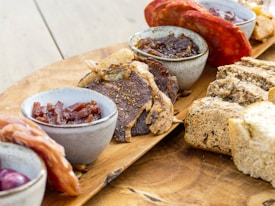 A wooden platter is arranged with a variety of cured meats, artisan breads, and small bowls containing different condiments. The presentation includes slices of dark bread, thinly sliced cured meats like salami and prosciutto, and thick pieces of beef jerky. The bowls appear to hold items like chutney and possibly pickled onions.