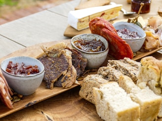 A wooden board displays an assortment of foods including various types of cold cuts, artisanal breads, and small bowls of preserves and pastes. Thinly sliced meats are arranged alongside dense, crusty bread. One bowl contains a reddish jam, another has a darker chutney, and a slice of brie cheese is visible in the background.