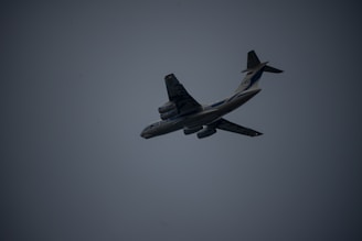 A large airplane is captured mid-flight against a dark, overcast sky. The aircraft is angled slightly upward, displaying its undercarriage and wing structure. The lighting is dim, creating a silhouette effect. The airplane features multiple engines and distinct markings on its body.