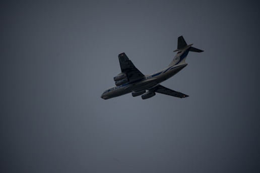 A large airplane is captured mid-flight against a dark, overcast sky. The aircraft is angled slightly upward, displaying its undercarriage and wing structure. The lighting is dim, creating a silhouette effect. The airplane features multiple engines and distinct markings on its body.