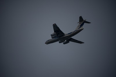 A large airplane is captured mid-flight against a dark, overcast sky. The aircraft is angled slightly upward, displaying its undercarriage and wing structure. The lighting is dim, creating a silhouette effect. The airplane features multiple engines and distinct markings on its body.