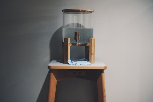 A transparent water dispenser sits on top of a wooden stand. The dispenser is partially filled with water and features a brass spigot. Below the spigot, a blue cloth is placed on the stand to catch any drips. The setup is against a plain gray wall, creating a minimalist appearance.