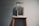 A transparent water dispenser sits on top of a wooden stand. The dispenser is partially filled with water and features a brass spigot. Below the spigot, a blue cloth is placed on the stand to catch any drips. The setup is against a plain gray wall, creating a minimalist appearance.