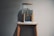 A transparent water dispenser sits on top of a wooden stand. The dispenser is partially filled with water and features a brass spigot. Below the spigot, a blue cloth is placed on the stand to catch any drips. The setup is against a plain gray wall, creating a minimalist appearance.