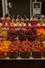 A market stall displays an array of fresh fruits including strawberries, grapes, kiwis, and oranges. In the background, there are cups filled with fruit pieces such as pineapple and watermelon, each with a straw sticking out. The setup is likely intended for takeaway fruit cups.
