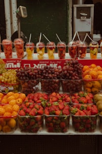 A market stall displays an array of fresh fruits including strawberries, grapes, kiwis, and oranges. In the background, there are cups filled with fruit pieces such as pineapple and watermelon, each with a straw sticking out. The setup is likely intended for takeaway fruit cups.