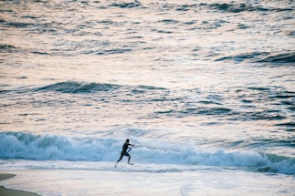 A person in a wetsuit runs along the shoreline, close to the breaking waves of an expansive ocean. The water appears slightly agitated with small waves and foam, and the lighting suggests late afternoon or early morning, casting a soft glow on the scene.
