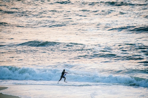 A person in a wetsuit runs along the shoreline, close to the breaking waves of an expansive ocean. The water appears slightly agitated with small waves and foam, and the lighting suggests late afternoon or early morning, casting a soft glow on the scene.