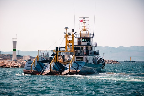 A ferry or transport vessel is seen moving away from the shore, with a small Turkish flag displayed on top. The vessel has large ramps, possibly for vehicles, and appears to be navigating through a calm sea. A lighthouse and rocky jetty are visible in the background, with distant mountains partly shrouded in mist.