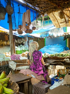 A vibrant marketplace scene with a variety of vegetables and fruits neatly arranged in baskets. An elderly woman with long gray hair, wearing a purple patterned sari, is seated amidst the produce, suggesting she might be a vendor. Above her, multiple hanging bags and cloths are visible, adding to the bustling atmosphere of the market. The setting is under a makeshift shelter composed of tarps and wooden planks, with natural light filtering through.