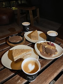 Three plates of desserts and three cups of coffee are arranged on a wooden table. Two slices of cake are visible; one layered with crumbs on top and another topped with chocolate and raspberries. Each cup of coffee has a heart-shaped design in the foam.