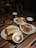 Three plates of desserts and three cups of coffee are arranged on a wooden table. Two slices of cake are visible; one layered with crumbs on top and another topped with chocolate and raspberries. Each cup of coffee has a heart-shaped design in the foam.