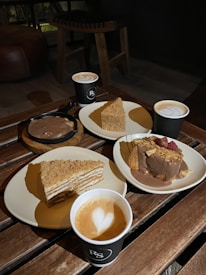 Three plates of desserts and three cups of coffee are arranged on a wooden table. Two slices of cake are visible; one layered with crumbs on top and another topped with chocolate and raspberries. Each cup of coffee has a heart-shaped design in the foam.