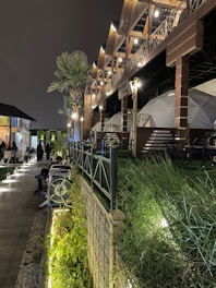 An outdoor setting featuring a modern patio with dome-shaped structures and seating arrangement surrounded by plants and trees. The area is illuminated with soft lighting from both the ground and overhead, giving it a cozy atmosphere. The structure has wooden elements and the night sky is visible.