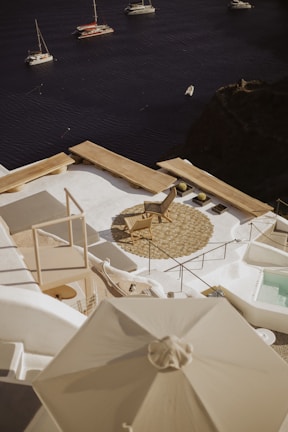 A serene coastal view featuring a luxurious outdoor terrace overlooking a calm sea. The terrace is adorned with a lounge chair, circular woven mat, and a white umbrella, all set against the backdrop of the deep blue water. Several sailboats float peacefully in the distance, adding to the tranquil ambiance.