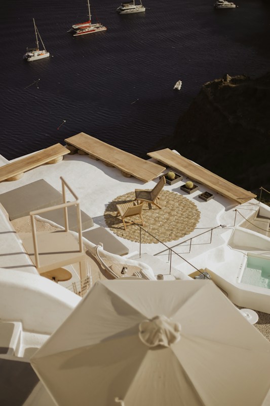 A serene coastal view featuring a luxurious outdoor terrace overlooking a calm sea. The terrace is adorned with a lounge chair, circular woven mat, and a white umbrella, all set against the backdrop of the deep blue water. Several sailboats float peacefully in the distance, adding to the tranquil ambiance.