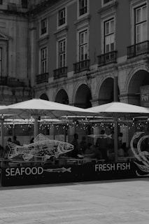 A black and white image of a building with arched windows and balconies. In front, there's an outdoor seating area with umbrellas and a sign displaying drawings of fish, labeled 'SEAFOOD FRESH FISH'. People are seated inside the enclosure, and string lights hang overhead.