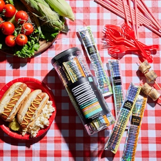 A vibrant picnic setup on a red and white checkered tablecloth, featuring a jar and individual packets of frozen cocktails labeled 'COOLOO'. Nearby, a variety of red plastic utensils is arranged, along with a bowl of tomatoes on lettuce leaves, sweet corn, and hot dogs with mustard on a plate alongside popcorn and pickles. Skewered roasted marshmallows add a sweet treat to the mix.