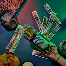 A tabletop arrangement with a variety of cocktail-flavored freeze pops and cocktail mixer accessories. In the background, a DJ controller and headphones suggest a music or party atmosphere. A bowl of colorful chips and dip are visible, and the setting is illuminated with vibrant lighting.