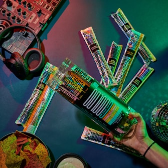 A tabletop arrangement with a variety of cocktail-flavored freeze pops and cocktail mixer accessories. In the background, a DJ controller and headphones suggest a music or party atmosphere. A bowl of colorful chips and dip are visible, and the setting is illuminated with vibrant lighting.