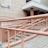 A beige and pink entrance ramp leads to a building, featuring multiple levels of handrails juxtaposed against a tiled walkway. The building has a light-colored tiled facade with a numbered sign and doorway slightly recessed under a covered entrance.