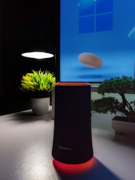 A modern still life setup on a desk featuring a black cylindrical speaker with a red glowing base in front of a digital blue screen displaying a 3D egg-like object. The scene is softly lit by a small desk lamp on the left, casting light onto a potted plant with green foliage. To the right, a small bonsai tree adds to the serene composition. The background screen contributes a futuristic ambiance to the setting.