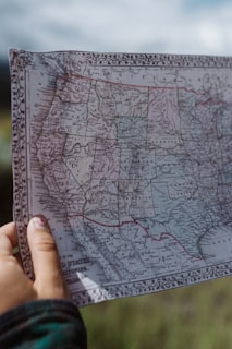 A hand holds up an old map of the western United States, featuring detailed outlines of states and territories. The map is bordered with an ornate design and shows labels of states such as California, Nevada, and Arizona. The sky is slightly overcast in the background.