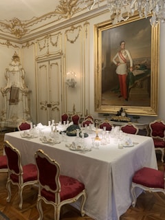 A lavishly decorated dining room featuring an opulent table setting with fine glassware and intricately folded napkins on a white tablecloth. The room is adorned with elaborate gold moldings and ornate furnishings, including red velvet chairs with gold trim. A large, formal portrait of a person in military-like attire hangs on the wall, surrounded by elegant wall decorations and sconces.