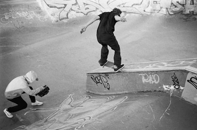A skateboarder in mid-action performing a trick on a ledge in a skatepark. The ground and walls are covered in graffiti, adding to the urban atmosphere. Another person is crouched down, holding a camera, capturing the moment. The image is in black and white, emphasizing the contrast and mood.