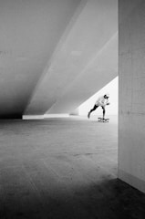 A black and white photograph featuring a skateboarder in motion under a large architectural structure. The scene is mostly empty, giving a sense of space and movement as the skateboarder glides through an expansive, open area. The lighting is dramatic, with strong contrasts between light and shadow accentuating the structural lines.
