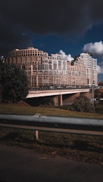 A large, modern apartment complex with an intricate geometric design comprises multiple connected buildings of varying heights. The architecture is highlighted by numerous windows and balconies. In the foreground, there's a grassy area with a metal railing, while dark storm clouds loom overhead, contrasting with patches of clear sky.