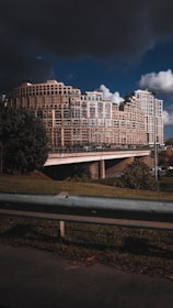 A large, modern apartment complex with an intricate geometric design comprises multiple connected buildings of varying heights. The architecture is highlighted by numerous windows and balconies. In the foreground, there's a grassy area with a metal railing, while dark storm clouds loom overhead, contrasting with patches of clear sky.