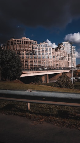 A large, modern apartment complex with an intricate geometric design comprises multiple connected buildings of varying heights. The architecture is highlighted by numerous windows and balconies. In the foreground, there's a grassy area with a metal railing, while dark storm clouds loom overhead, contrasting with patches of clear sky.