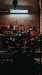 A dimly lit workshop with a focus on a vintage motorcycle surrounded by various tools and equipment. A fluorescent light hangs overhead, casting a bluish glow on the scene. The shelves in the background hold an assortment of items, including books and mechanical parts.
