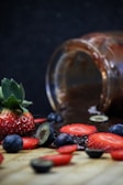 A glass jar lies on its side with dark liquid spilling out, surrounded by fresh strawberries and blueberries on a wooden surface. Some berries are sliced, displaying their vibrant colors against a dark background.