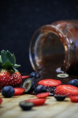A glass jar lies on its side with dark liquid spilling out, surrounded by fresh strawberries and blueberries on a wooden surface. Some berries are sliced, displaying their vibrant colors against a dark background.