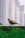 A bird with speckled plumage is perched on a wooden fence. In the background, a series of tall, blurred buildings create an urban setting. The focus is sharp on the bird, with some greenery in the foreground adding a natural element.