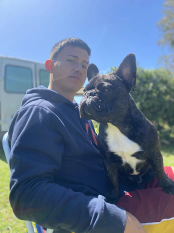 A close-up of a smiling dog owner cuddling their French Bulldog outside on a sunny day.