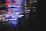 A moody photograph of city lights reflected on wet pavement with a purple and cyan tint.
