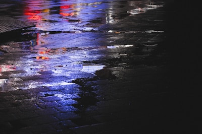 A moody photograph of city lights reflected on wet pavement with a purple and cyan tint.