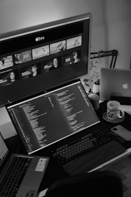 A monochrome setup with a large monitor displaying Apple TV+ content and a secondary monitor showing code. Two laptops, a cup, a smartphone, and a keyboard are also present on the desk.