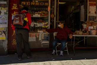 A person with a backpack stands outside a bakery, interacting with another person seated at a table, who is wearing a mask. The storefront features various signs, while baked goods and other items are displayed on the table.