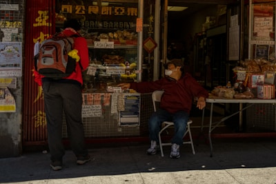 A person with a backpack stands outside a bakery, interacting with another person seated at a table, who is wearing a mask. The storefront features various signs, while baked goods and other items are displayed on the table.