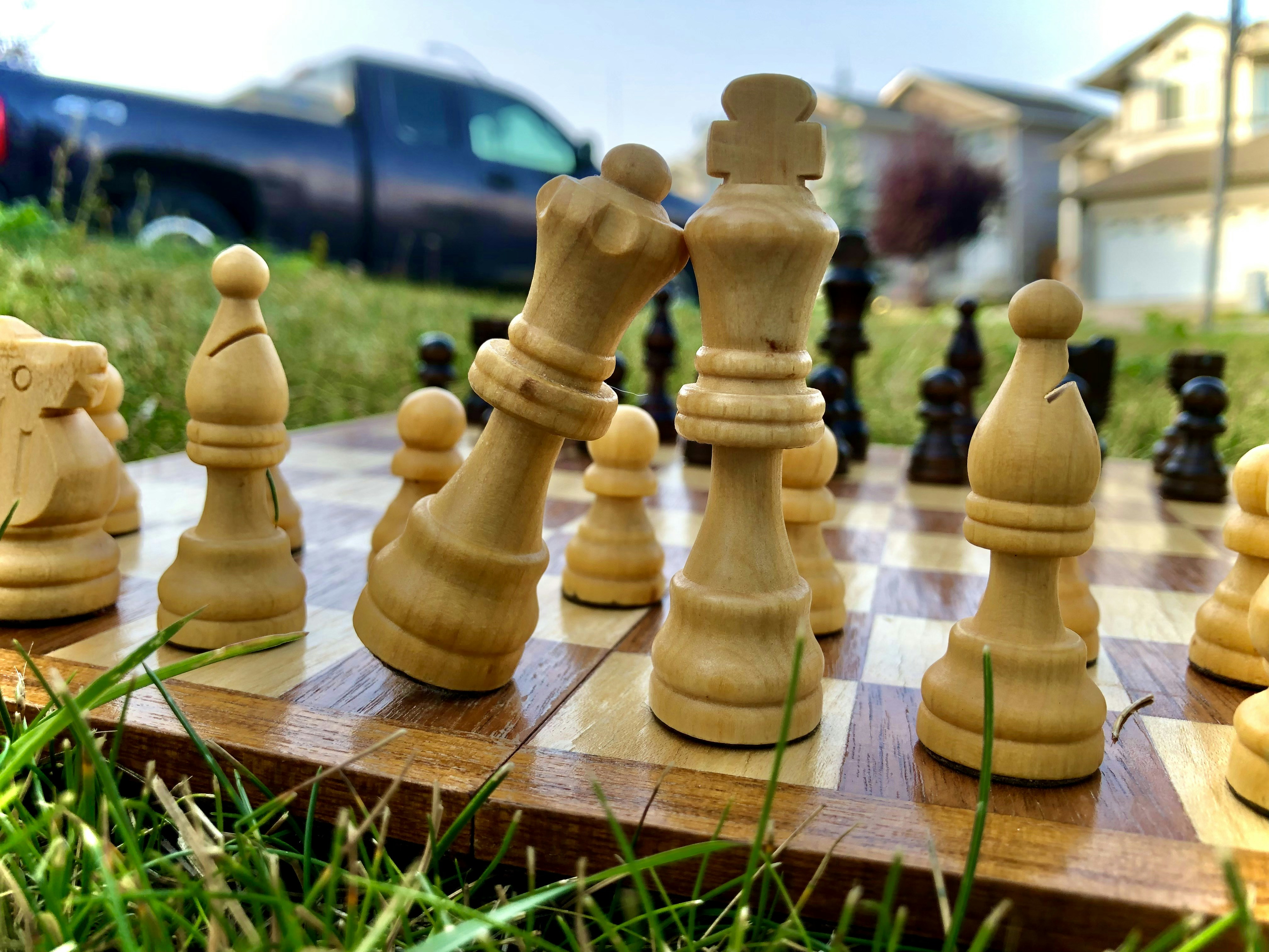 Chess pieces arranged on a board placed outdoors, with grass in the foreground. The image features a blurred background with a truck and residential buildings.